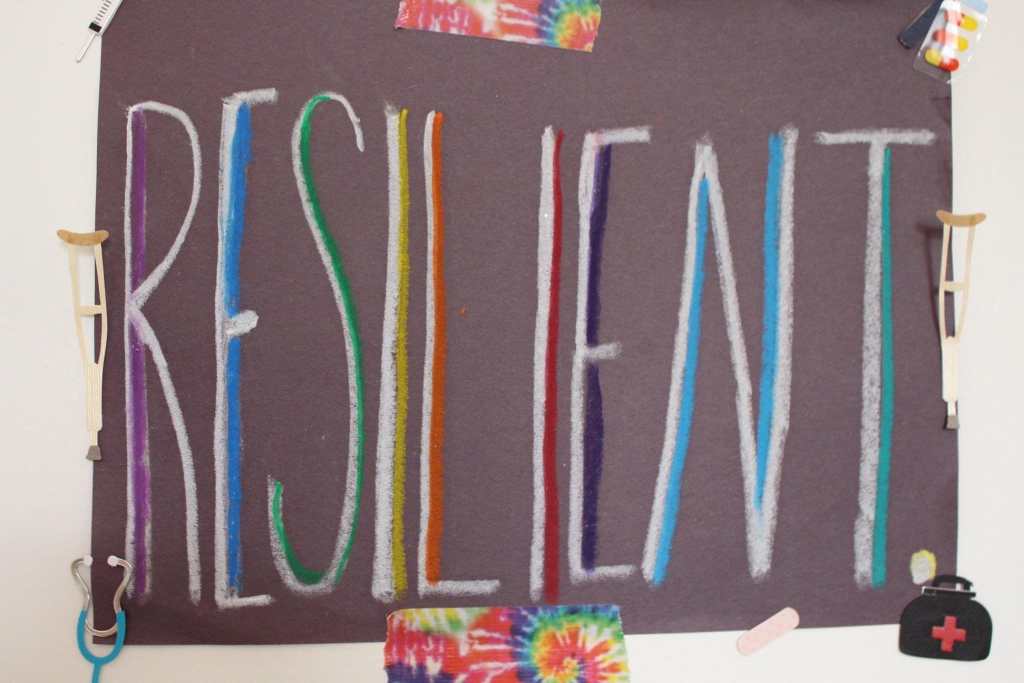 [RESILIENT in rainbow Attached to the wall with stickers of crutches, a stethoscope, a bandaid, a first aid kid, medicine, and rainbow tie dye tape.]