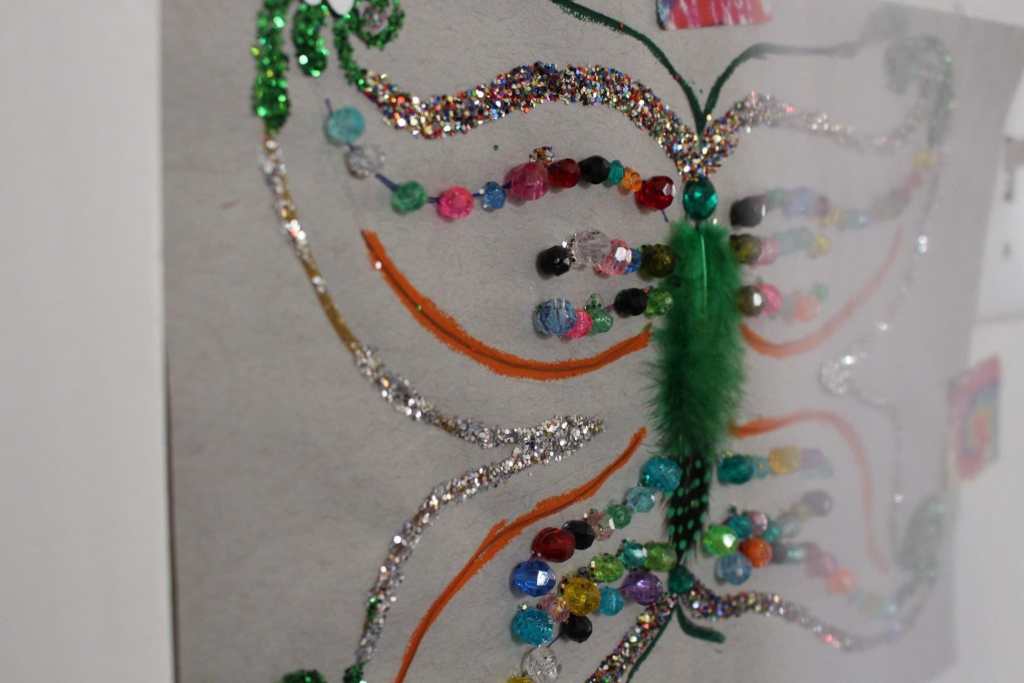 [A rainbow butterfly with a green feather body, silver glitter wings, rainbow beaded wings, gold and glitter wings, on gray paper.]