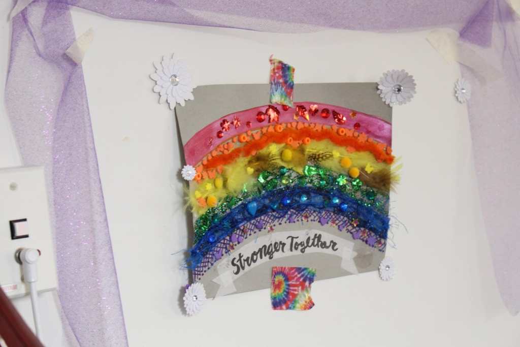 [Purple tulle ribbon draped around a piece of art. The piece of art is a rainbow made of various fabrics and materials, on gray paper. Under the rainbow are the words “Stronger Together” in black cursive handwriting on white background. It is attached to the wall with white flower stickers and tie dye rainbow tape.]