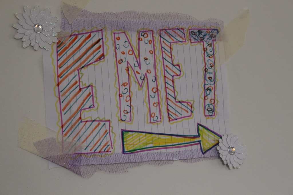 [A sign saying EMET, in block letters with patterns, like stripes, and polka dots, inside them. Below it is a yellow arrow pointing to the right, indicating Emet. It is attached to the wall with white flower stickers.]