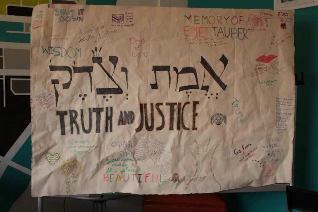 [A white canvass banner with Emet (Truth) & Justice written in Hebrew calligraphy: אמת צדת Around it are notes from those who loved Emet, in multicolored print.]