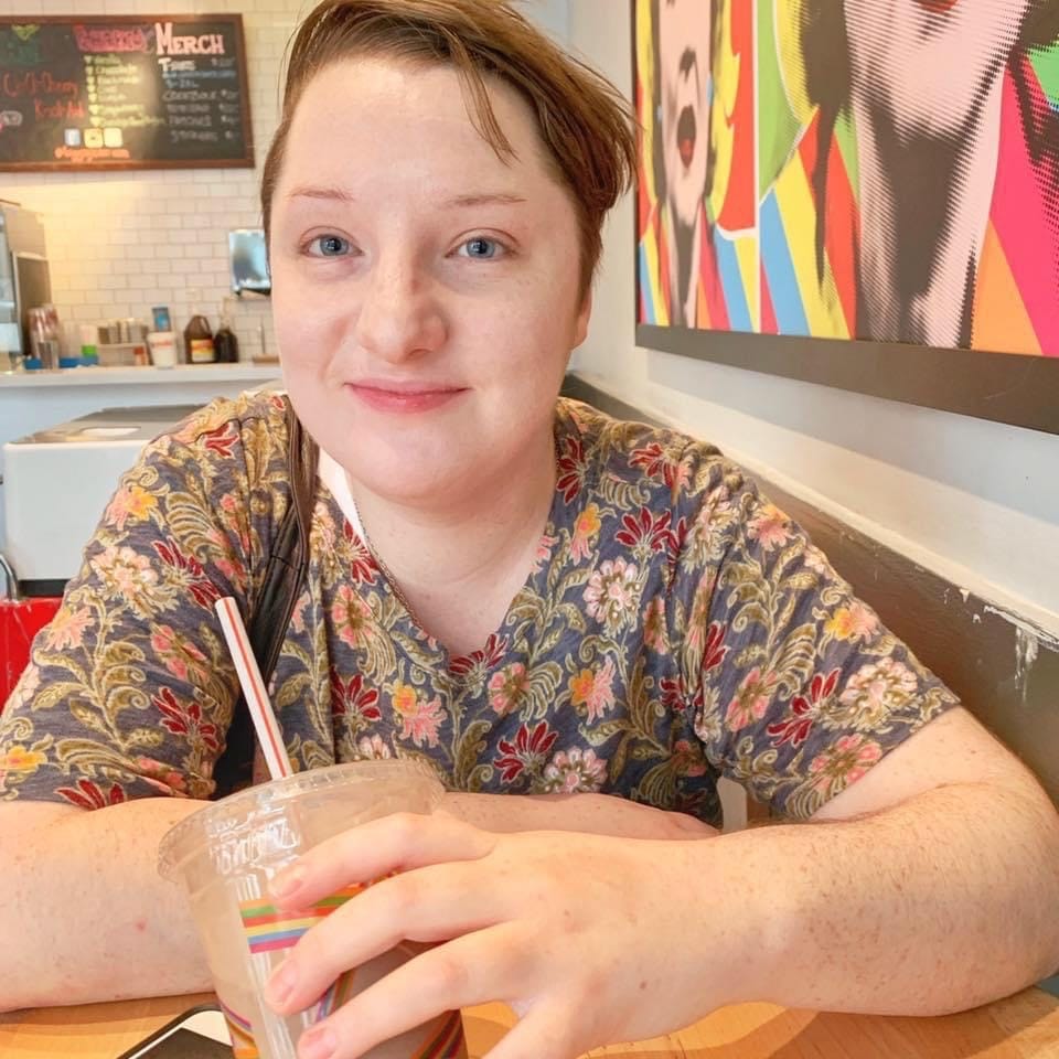 Noah, a white trans masc non-binary person with short brown hair, smiles at the camera while holding a milkshake. They are wearing a short sleeved green shirt with red, orange, and pink flower pattern on it.