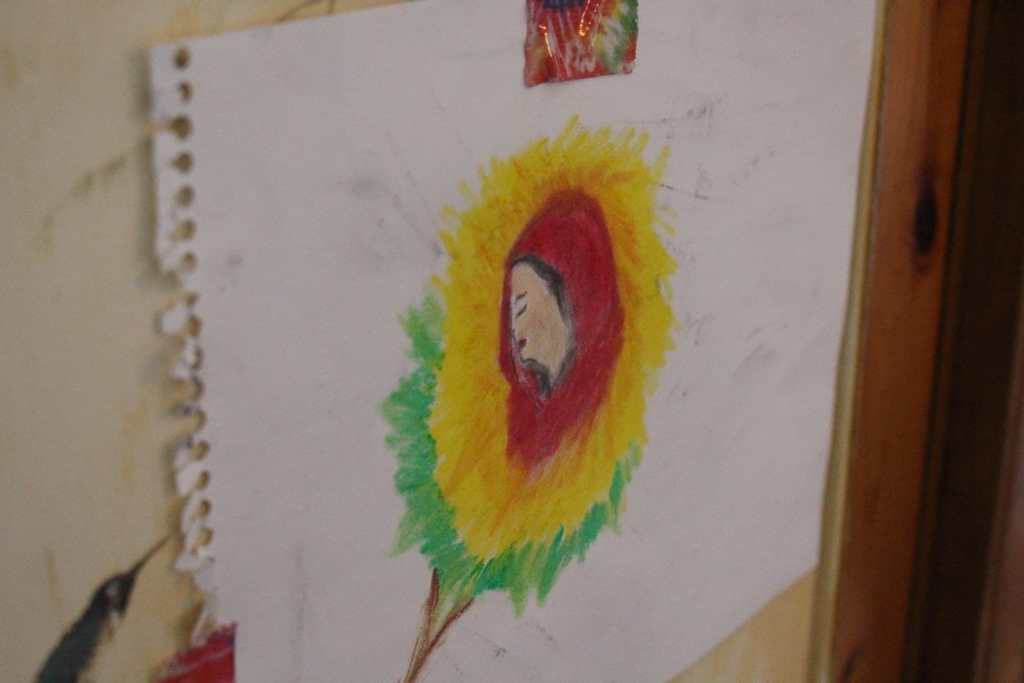 [A piece of art on white paper depicting a woman wearing a red head covering, and surrounding her are yellow and green rings of light. It is attached to the wall with rainbow tie dye tape.]