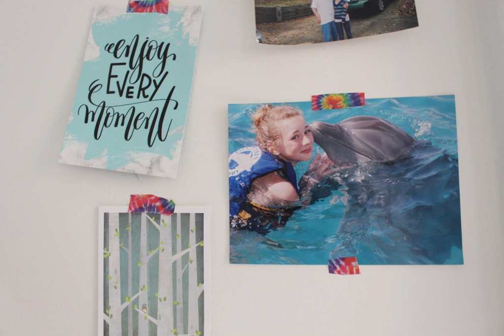 [A childhood picture of Emet’s sister, Rachel, swimming with a dolphin which appears to be nuzzling her. She is wearing a life vest and smiling. It is attached to the wall with rainbow tie dye tape. Next to it is a card with the words “enjoy every moment.” It is attached to the wall with rainbow tie dye tape. Below is a card depicting trees, also attached to the wall with rainbow tie dye tape.]