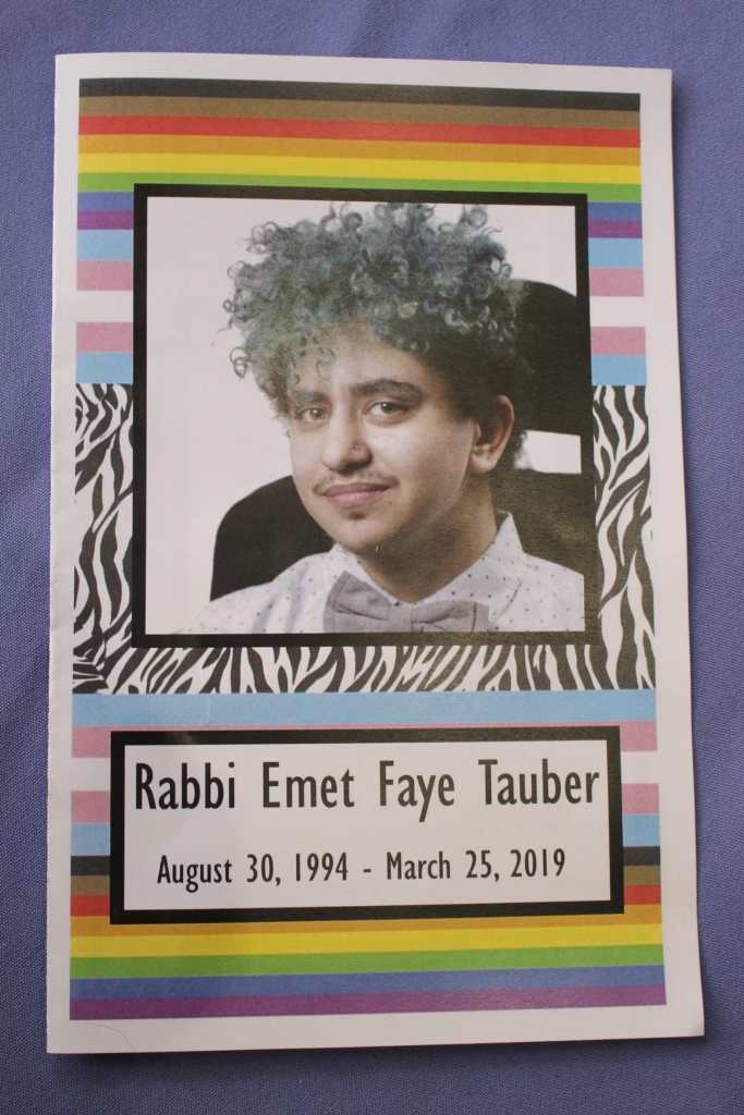 [Emet’s funeral program. On the cover is a picture of Emet wearing a white and gray polka dotted collared button up shirt, with a gray bow tie. He has curly turquoise hair. He has a mustache and a nose piercing and is sitting in a power chair, smiling. Below it says Rabbi Emet Faye Tauber August 30, 1994-March 25, 2019 In the background there are zebra stripes, rainbow pride flags, and trans pride flags.]