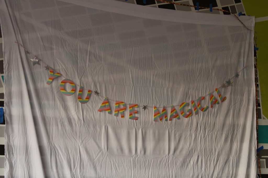 [A garland that says “YOU ARE MAGICAL” in rainbow striped letters, draped over a white sheet.]