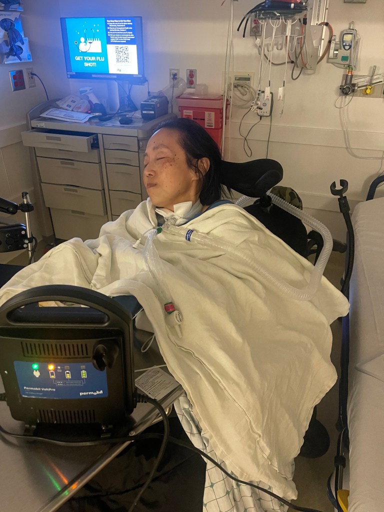 A picture of me, an Asian American disabled woman in a wheelchair, a tracheostomy is in my throat connected to a ventilator tube. I am in the emergency room at UCSF Moffitt/Long hospital. I am swaddled in blankets, my eyes are closed, and I am trying to sleep as I am waiting for test results and a bed in the intensive care unit. Behind me is a computer monitor with a bright glow. On a table in front of me is a wheelchair battery charger.
