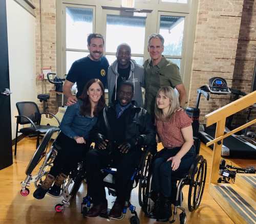 Group photo of 6 people from a shoot on NCIS: New Orleans. In the front row from left to right are three wheelchair users composed of two white women and one Black man: Katherine Beattie, Daryl Chill Mitchell, and Teal Sherer. In the back are three white men standing left to right: Kurt Yeager, LeVar Burton, and Scott Bakula. Photo courtesy of Katherine Beattie.