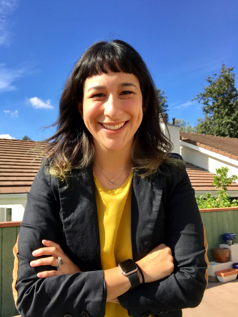 A medium-skinned person with black hair worn long around their shoulders and short baby bangs, smiling outdoors, with a blue sky in the background.