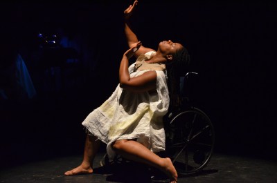 Image of a self-described, 'an African American, queer, disabled, femme' sitting in a manual wheelchair. Her head is tilted back, arms and hands raised upward, her legs are resting on the ground at different angle, one leg straight while the other is bent 45 degrees. The background is completely dark.