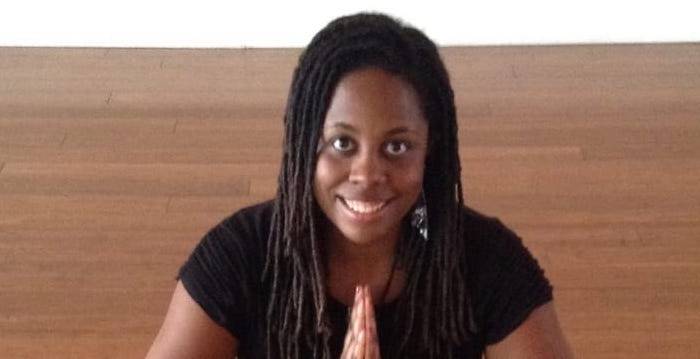 IMAGE OF MIXED ABILITIES DANCE TEACHER INDIA HARVILLE. SHE IS AFRICAN-AMERICAN WITH HAIR IN LOCKS, WEARING ALL BLACK, SITTING ON THE FLOOR WITH HER HANDS CLASPED, SMILING AT THE CAMERA.