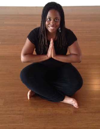 IMAGE OF MIXED ABILITIES DANCE TEACHER INDIA HARVILLE. SHE IS AFRICAN-AMERICAN WITH HAIR IN LOCKS, WEARING ALL BLACK, SITTING ON THE FLOOR WITH HER HANDS CLASPED, SMILING AT THE CAMERA.