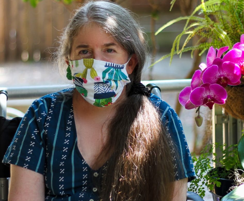 Photo of Ingrid, a middle-aged White woman with a long gray/brown ponytail who is wearing a bright blue dress and a colorful cloth face-mask. She's sitting in her wheelchair outdoors, next to a fuschia orchid. Photo credit: Christopher Egusa