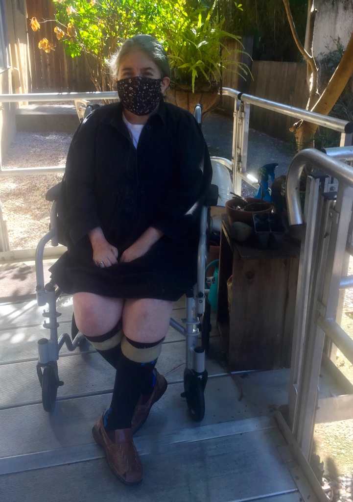 Five weeks later. The same woman, Ingrid Tischer in a cloth mask and shoe orthotics sitting outside in a transport chair on a ramp in the sun.
