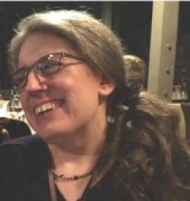 Image of an older white woman with glasses, her long hair is puled back and resting on one shoulder. She is looking away from the camera and smiling. She is wearing a dark-colored shirt and a necklace.