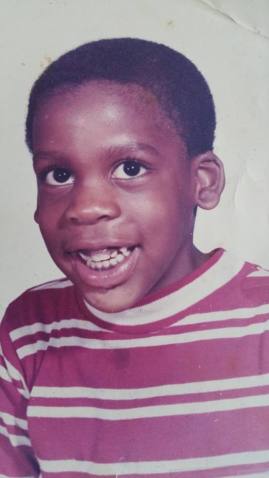 A young Black boy, Leroy Moore, with short black hair smiling at the camera. He is wearing a red and white striped shirt.
