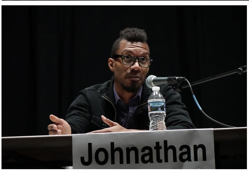 Johnathan, a black man with glasses, is seated at a table behind a microphone. He appears to be using his hands to emphasize his explanation, and his expression is one of challenge. A placard with his name on it and a bottle of water are also on the table.