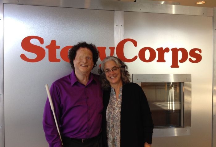 Picture of two individuals standing in front of a StoryCorps recording booth. A man is on the left side wearing a purple shirt and black pants. He is holding a cane The woman next to him has long white and dark hair and wears glasses. She is wearing a black cardigan sweater. Both are smiling at the camera.