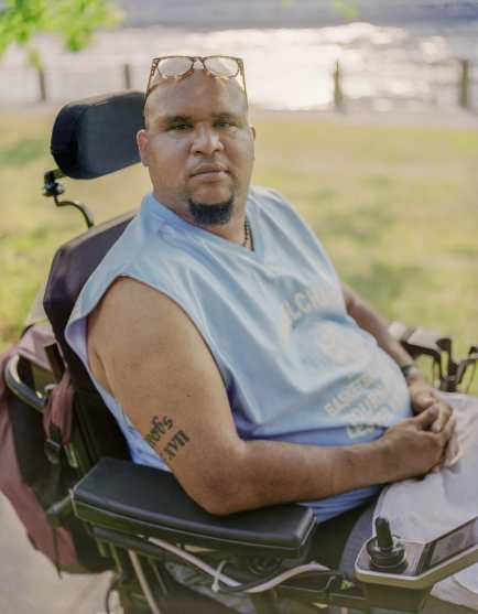 Jay, a Dominican man with a goatee and reading glasses perched on top of his head, sits in a wheelchair outside on a sunny day. There is grass and water in the background. He wears a light blue T-shirt and looks straight into the camera with a serious expression. Photo credit: Elias Williams
