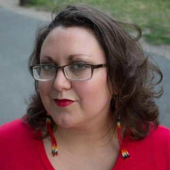Close up of a light complexion, Two Spirit femme with black glasses, green eyes, shoulder length brown hair, Native style beaded dangling earrings, with bright red lips and a red shirt. In the foreground is a paved path and grass.