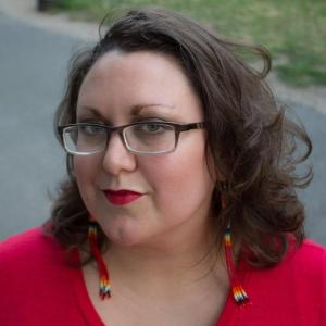 Close up of a light complexion, Two Spirit femme with black glasses, green eyes, shoulder length brown hair, Native style beaded dangling earrings, with bright red lips and a red shirt. In the foreground is a paved path and grass.