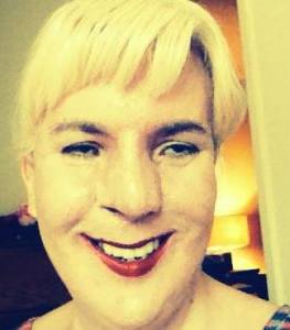 Image of Jennifer Justice, a white woman with short blonde hair. She is wearing bright red lipstick and is smiling at the camera. Her image has a filter that gives a yellow tint.  