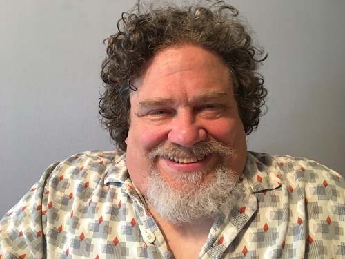 A middle-aged man smiles, He has brown curly hair, a grey goatee and wears a vintage looking cocktail hour shirt.