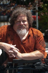 Photo of a man in a wheelchair, turned so his arms are resting on one side of the wheelchair's armrest. He is an older white man with brown curly hair and a white beard. It is a sunny day and he is wearing a short sleeved orange shirt with a pattern.