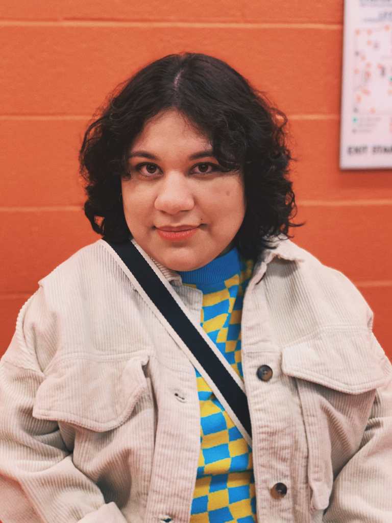 Photo of Jocelyn Mondragon, a Mexican woman with curly short black hair, smiling straight at the camera. In front of an orange painted brick wall, Jocelyn is wearing a beige corduroy jacket with a blue and yellow checkered sweater underneath. A black and white striped strap is across her shoulder.