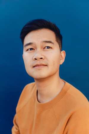 Jonny Sun, a young Chinese Canadian man with short black hair wearing an orange crewneck shirt. He is looking upward at the sky pensively. There is a deep blue background behind him. Photo credit: Rozette Rago
