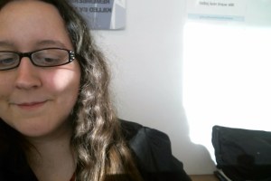 A young white woman on the left-side of the image looking downward. She has long wavy brown hair and eyeglasses on. On the right is a bright spot of sunlight.