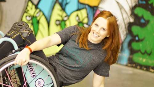 Katherine Beattie, a young white woman with long red hair. She is wearing a gray t-shirt and in her wheelchair popped at a 45 degree angle against a colorful mural.