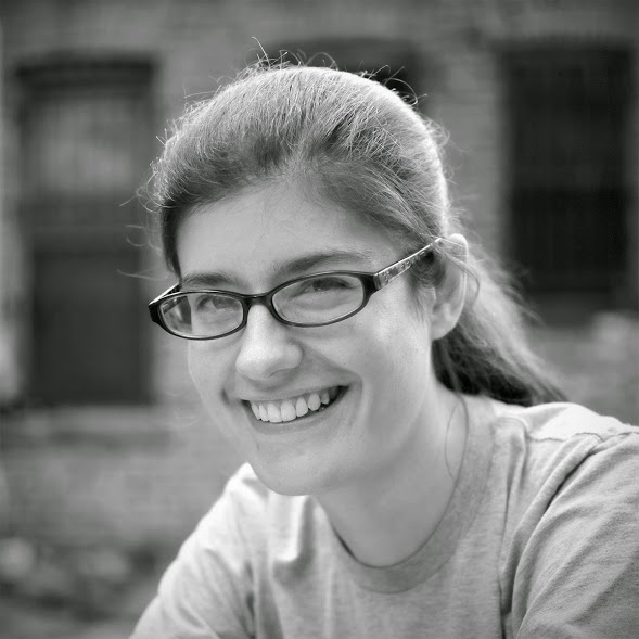 A black-and-white headshot photo of Katherine, a white woman with long brown hair pulled back wearing oval plastic-rimmed glasses and a grey T-shirt, smiling at the camera