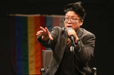 A brown round queer with glasses and short spiky hair performs at a microphone in front of the rainbow and transgender flags. They smile holding a microphone with their hand outstretched.