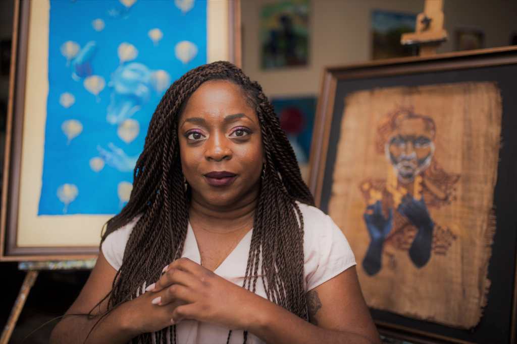 Photo of Kemi Yemi-Ese, a young Nigerian American Black woman with long black braids. She is wearing a white short-sleeved shirt and her hands are clasped in front of her. Behind her are two canvases featuring her paintings, the one on the left is light blue painting with abstract shapes and the one on the right is a painting of an elder Black man 