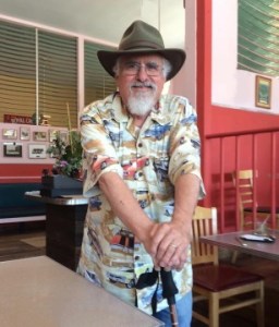Older white man with a fedora-type hat and a short-sleeve printed shirt. He is standing and both of his hands are resting on top of his cane. He has white hair and a white beard. He is standing inside a cafe.