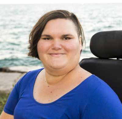 A headshot of Kennedy, a white, fat, femme, smiling softly at the camera. She has chin length brown hair with one side shaved and is wearing a blue shirt. Her electric wheelchair and Lake Michigan are behind her. 