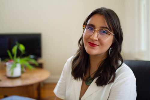 Photo of Kera Sherwood-O'Regan, an Indigenous Māori woman with light olive skin and long brown hair, sitting in her living room. She is wearing a white collared jumpsuit, vintage style tortoiseshell glasses, Haus of Dizzy love heart earrings, and red lipstick. She wears a Māori pounamu greenstone pendant around her neck, and she is smiling slightly at the camera. There is a green houseplant and wooden furniture in the background.