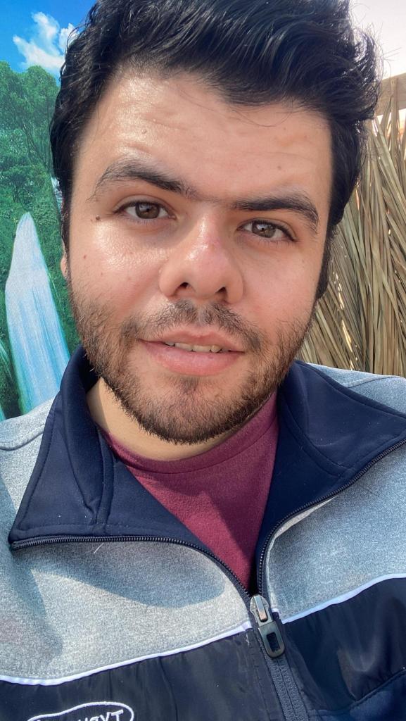 close-up of Khaled outside in the sun, dark brown hair combed up, slight brown beard and hazel-brown eyes staring into the camera. He wears a slight smile and a blue and gray zip up with maroon shirt underneath. 