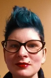 Young white woman with blue hair styled upward like a mohawk (but not). She has cat-eyed eyeglasses on and bright red lipstick. 