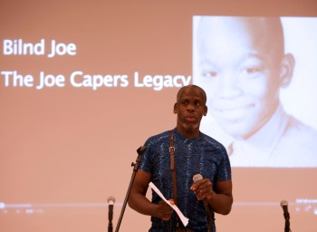 Image description: Leroy F. Moore, Jr., a Black disabled man giving a lecture. He is standing in front of a microphone and wearing a short-sleeve blue shirt. Behind him on a screen is a slide showing a black and white photo of a young Black child. Left of that photo is text that reads, "Blind Joe, The Joe Capers Legacy."