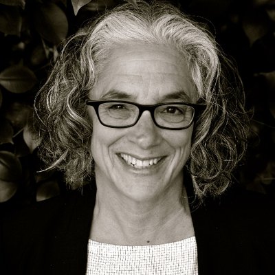 A black-and white photo of an older white woman with dark-colored eyeglass frames, shoulder-length curly hair that's white and gray. She is standing behind a dark background wearing a black blazer and a white scoop neck top. She is smiling at the camera. Photo credit: Ahri Golden