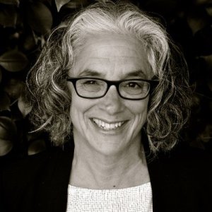 A black-and white photo of an older white woman with dark-colored eyeglass frames, shoulder-length curly hair that's white and gray. She is standing behind a dark background wearing a black blazer and a white scoop neck top. She is smiling at the camera. Photo credit: Ahri Golden
