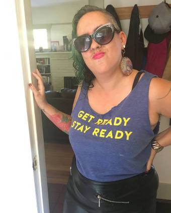 Image description: Leah, a light brown skinned femme in her early 40s, faces the camera with her hand on her right hip. She wears big oversigned vintage sunglasses with rhinestones, a blue t shirt that says "Get Ready, Stay Ready" in yellow lettering, and a black leather skirt. She has hot magenta lipstick, brown and green curly hair that is shaved on one side, and one large round earring with an image of Frida Kahlo is visible on her right side.