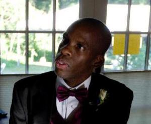 Image of Leroy Moore, a Black man with a shaved head looking left from the camera. He is wearing a black tuxedo with white shirt and magenta bow tie. Behind him is a glass-paned window.