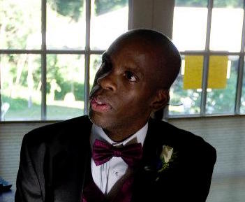Image of Leroy Moore, a Black man with a shaved head looking left from the camera. He is wearing a black tuxedo with white shirt and magenta bow tie. Behind him is a glass-paned window.