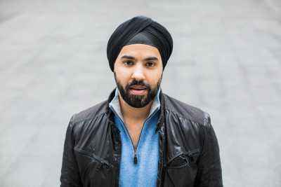 Sikh man wearing a black turban, a black leather jacket and a light blue shirt underneath. He has a beard with black hair and standing outdoors with a gray background. Photo credit: Les Talusan