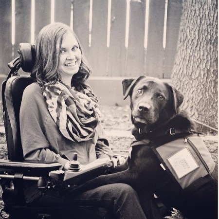 Young white woman in a sepia-tone photo. She is in a power chair and a black labrador retriever that is her service animal has its two front paws on her lap.