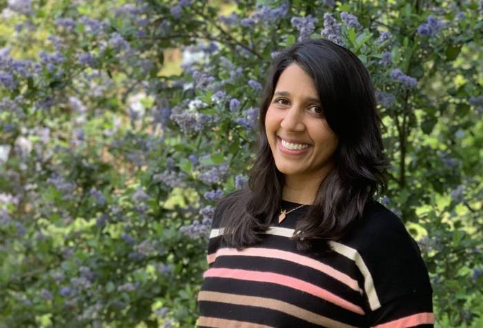 An Indian woman wearing a black sweater with pink and orange stripes and blue jeans smiles in front of a green tree with purple flowers. Her hands are in her pockets.