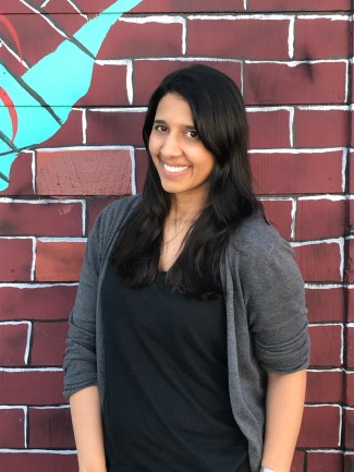 Image description: smiling Indian woman wearing jeans, a black shirt, and grey cardigan standing in front of a painted brick wall.
